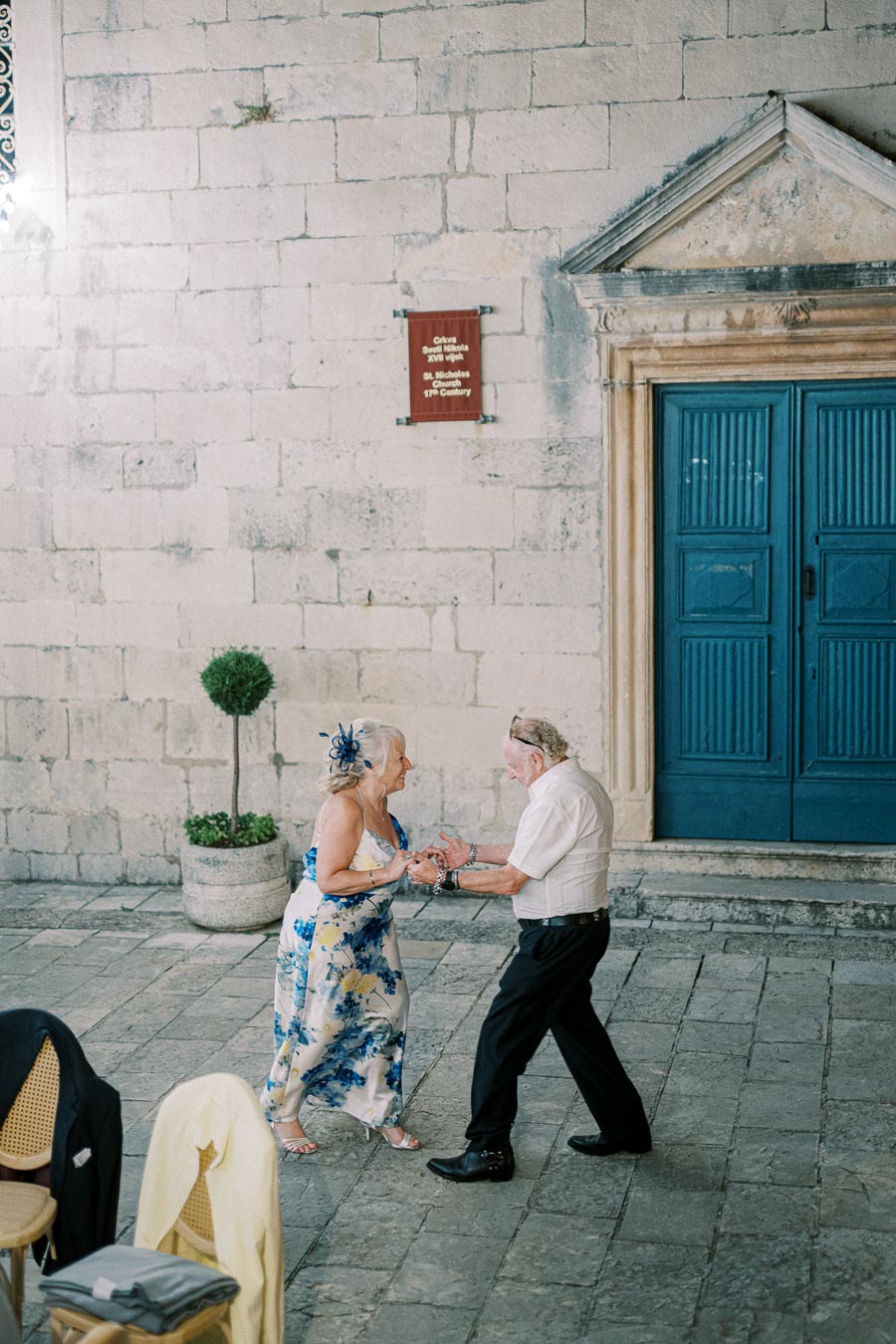 Elderly couple dancing joyfully outside a historic stone building with blue doors, showcasing love and vitality in an outdoor setting.