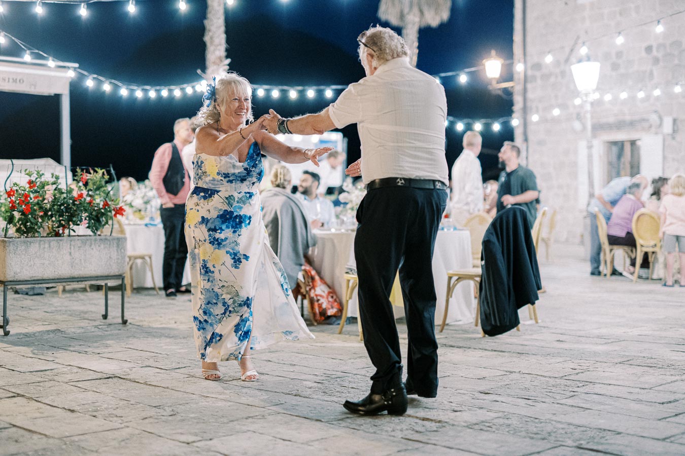 Elderly couple dancing joyfully at a nighttime outdoor celebration, surrounded by festive string lights and guests seated at tables.
