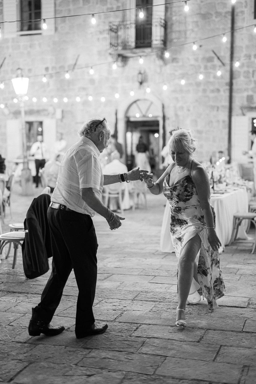 Elderly couple dancing joyfully at an outdoor evening event, surrounded by string lights and a historic stone building backdrop.