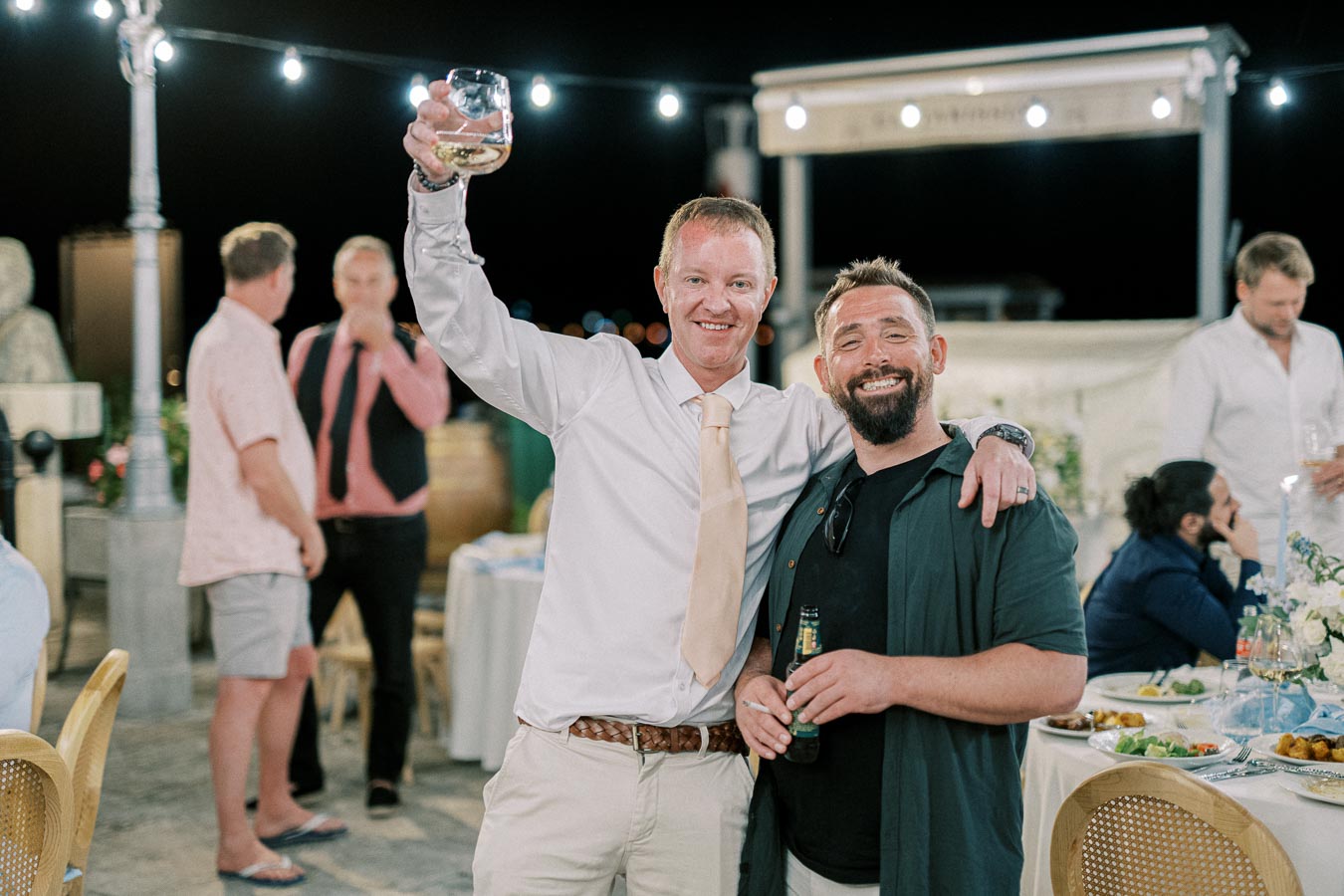 Two smiling men celebrating at an outdoor evening event, one holding a glass of wine and the other a bottle of beer, with string lights overhead and a festive atmosphere.