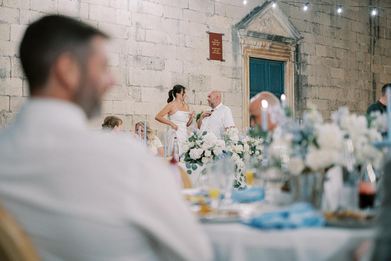 Wedding reception scene with a bride and groom in conversation, adorned dining table in foreground, rustic stone wall and string lights in background, creating a romantic atmosphere.