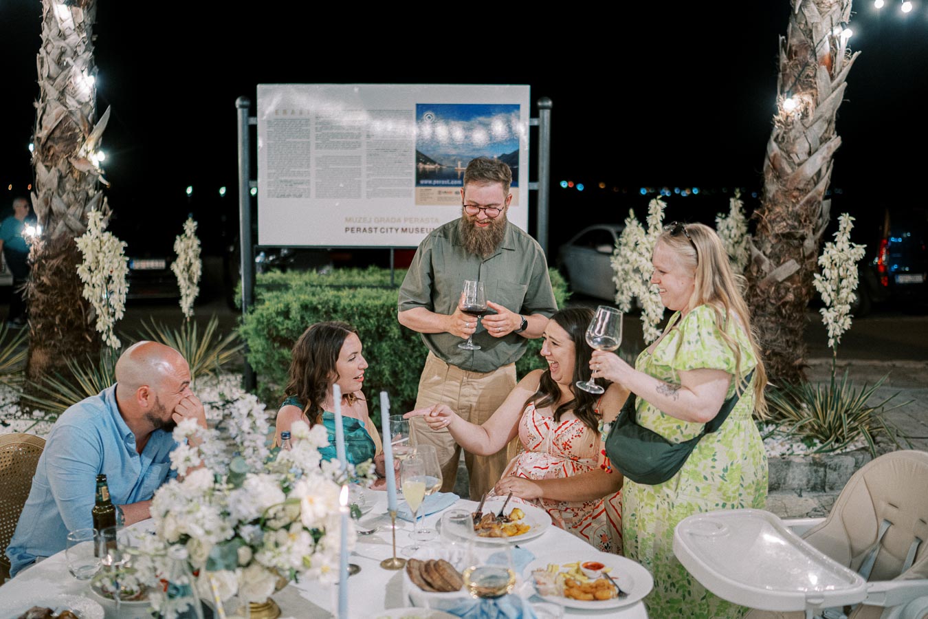 Group of people enjoying an outdoor evening gathering with wine and food at a decorated table under palm trees.