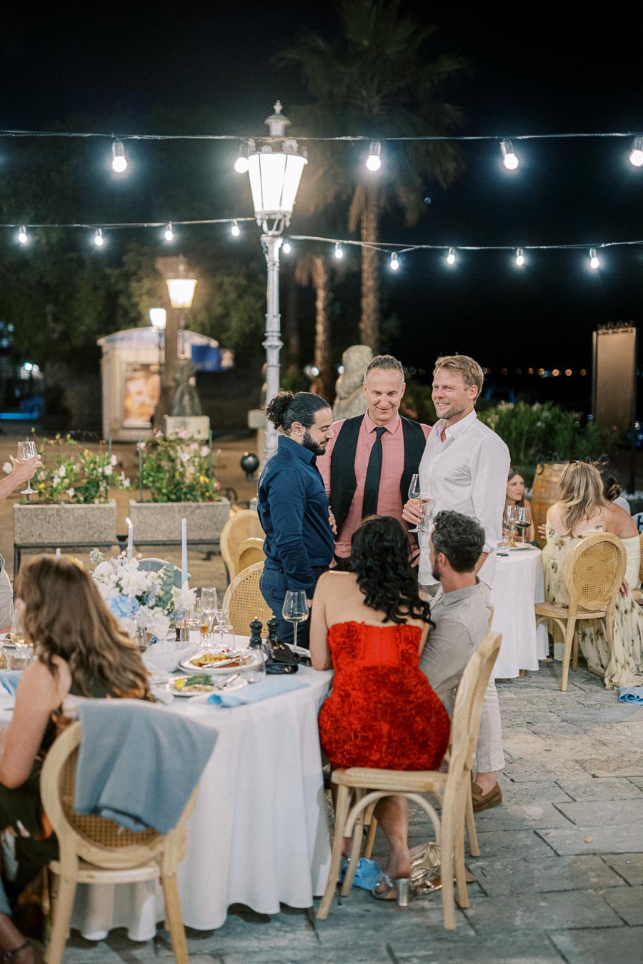 Outdoor evening gathering with elegantly dressed guests socializing around a candlelit table, under string lights and street lamps, creating a festive atmosphere.