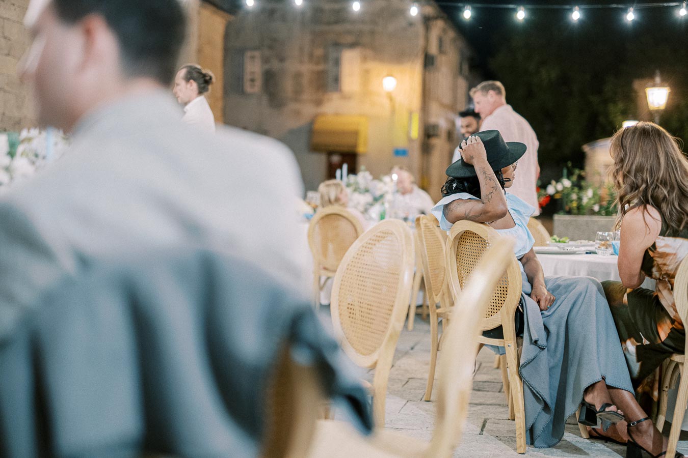 Outdoor evening gathering with people seated at tables under string lights. The focus is on a person in a hat and blue dress, engaged in conversation. The setting has a warm, inviting atmosphere with rustic stone walls and ambient lighting.