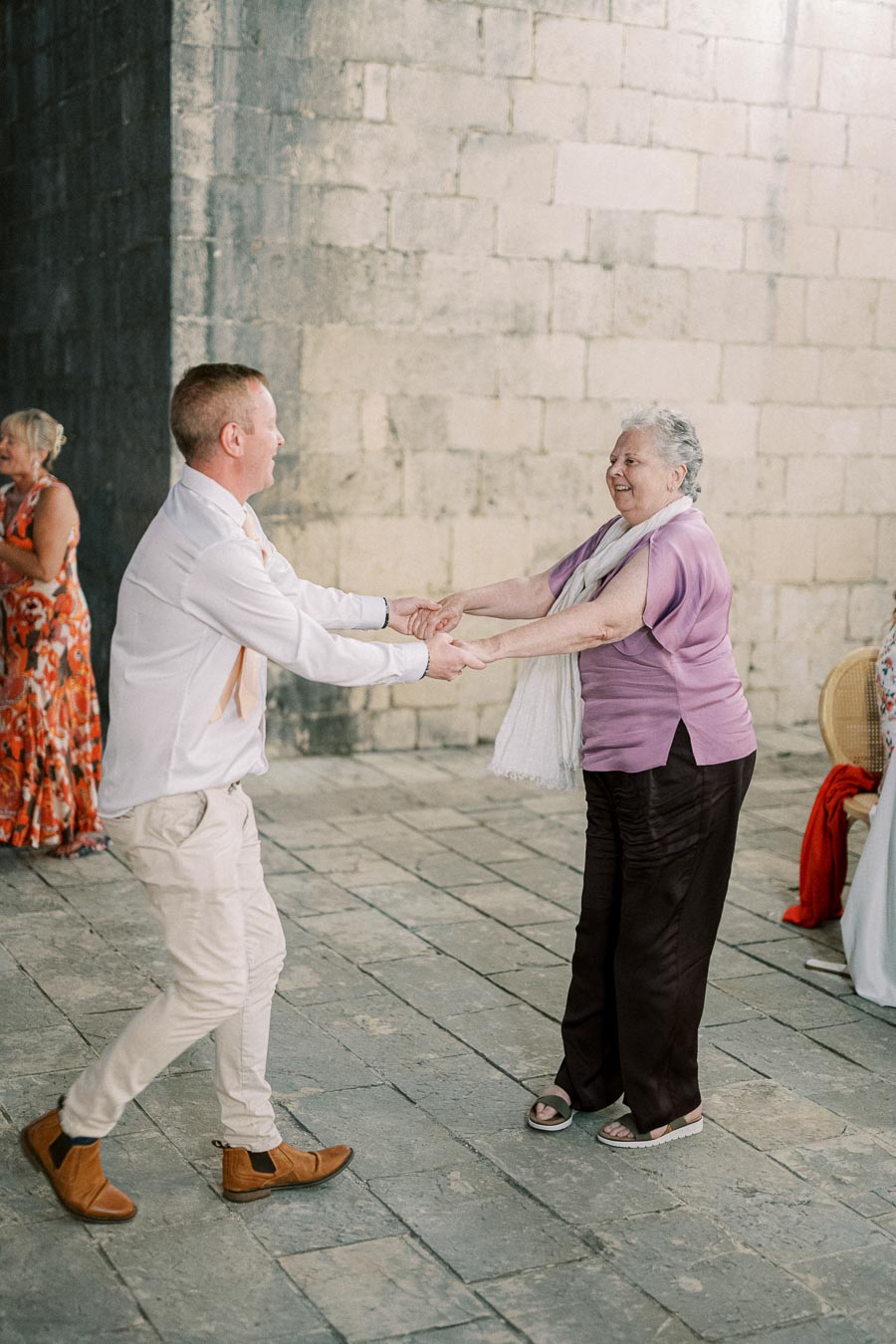Man in white shirt dancing with elderly woman in a purple blouse, sharing a joyful moment at a celebration event with stone wall background
