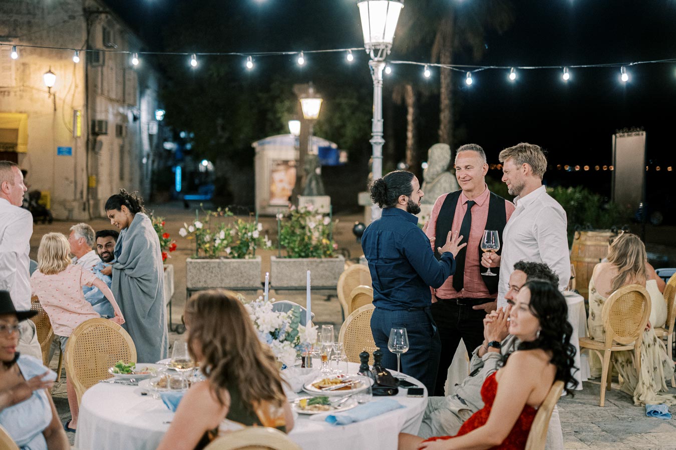 Outdoor evening gathering with people socializing at a wedding reception. Guests sit and stand around tables with food and wine glasses, under string lights creating a festive atmosphere.