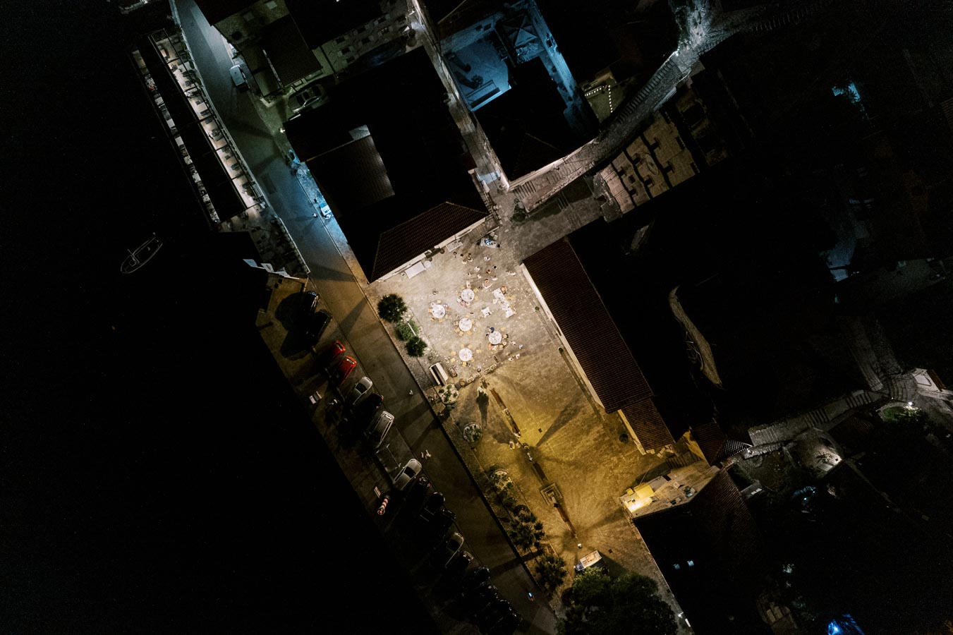 Aerial night view of an illuminated urban courtyard surrounded by buildings, with tables and chairs arranged in the center, casting shadows under streetlights.