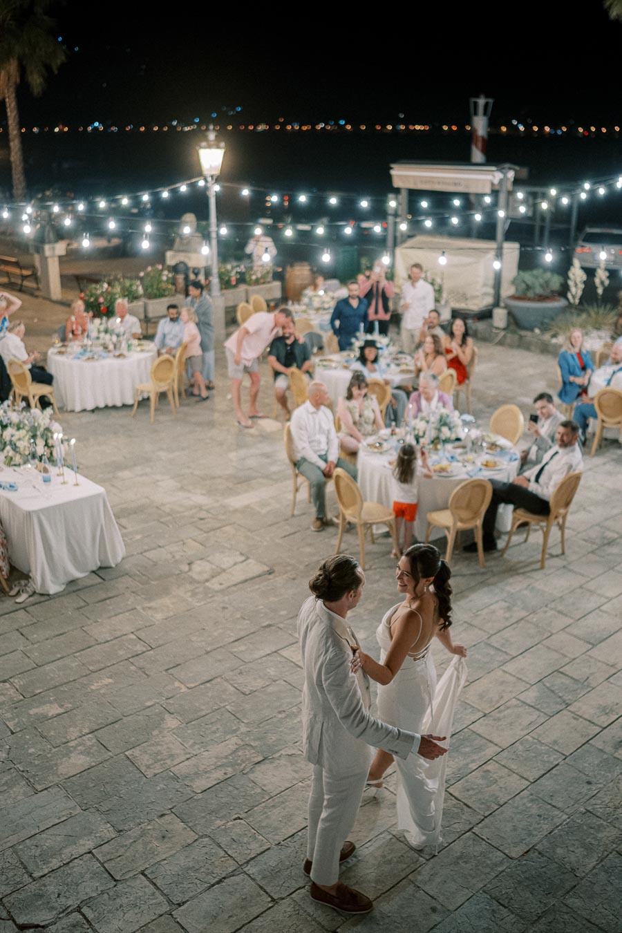 A couple shares their first dance at an outdoor wedding reception, surrounded by seated guests under string lights on a stone patio.