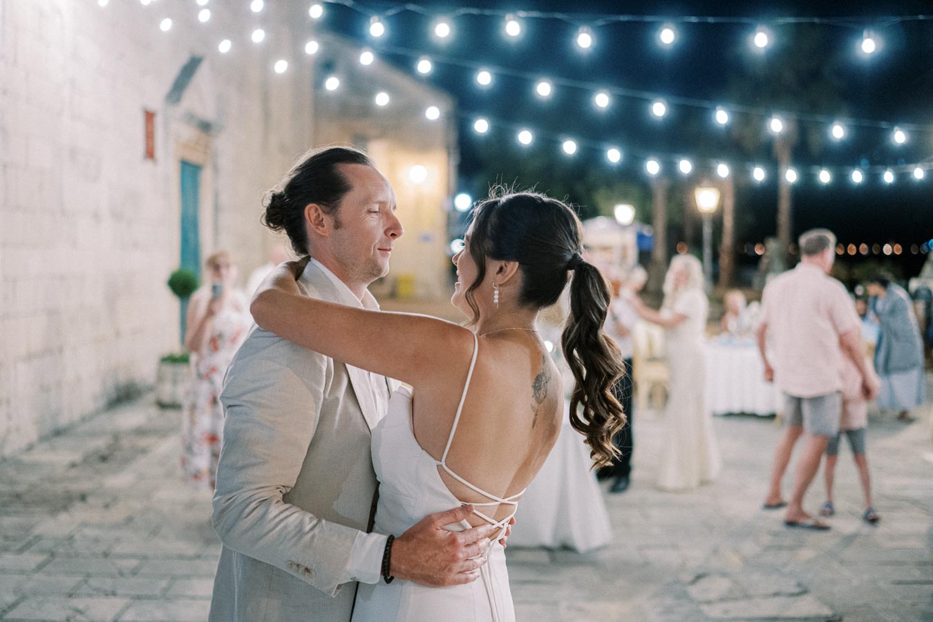 A couple sharing a romantic dance at an outdoor wedding reception, surrounded by string lights and guests enjoying the evening ambiance.