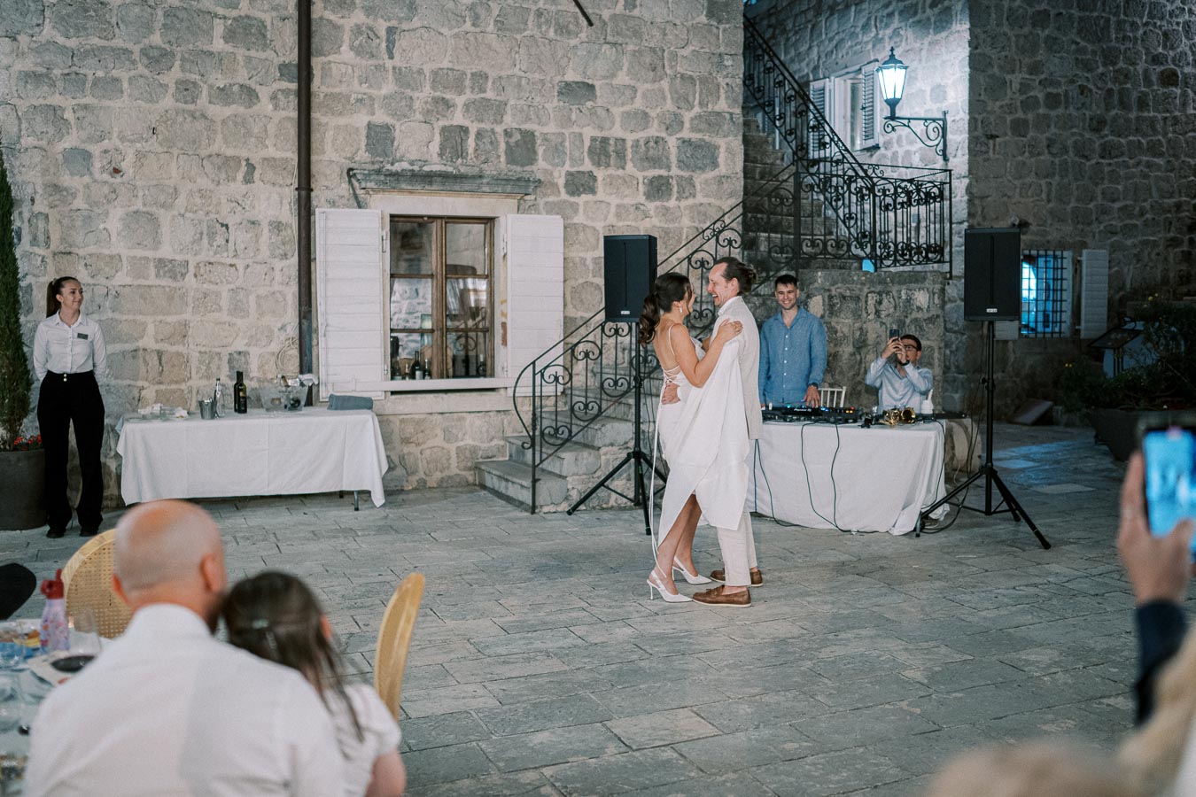 Couple dancing at an outdoor wedding reception with stone building backdrop and DJ setup.
