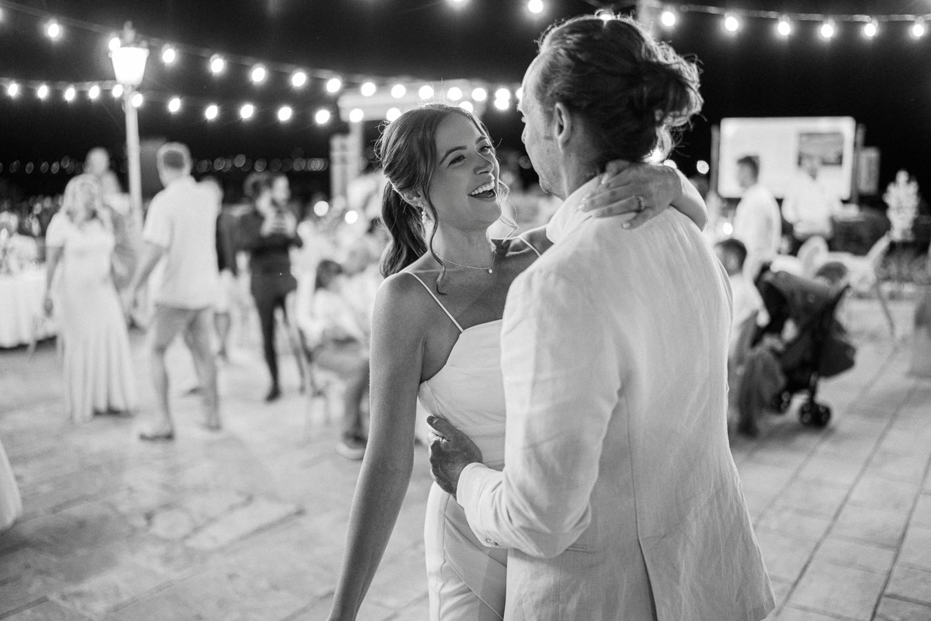 A couple dances joyfully under string lights during an outdoor evening celebration, surrounded by guests at a party.