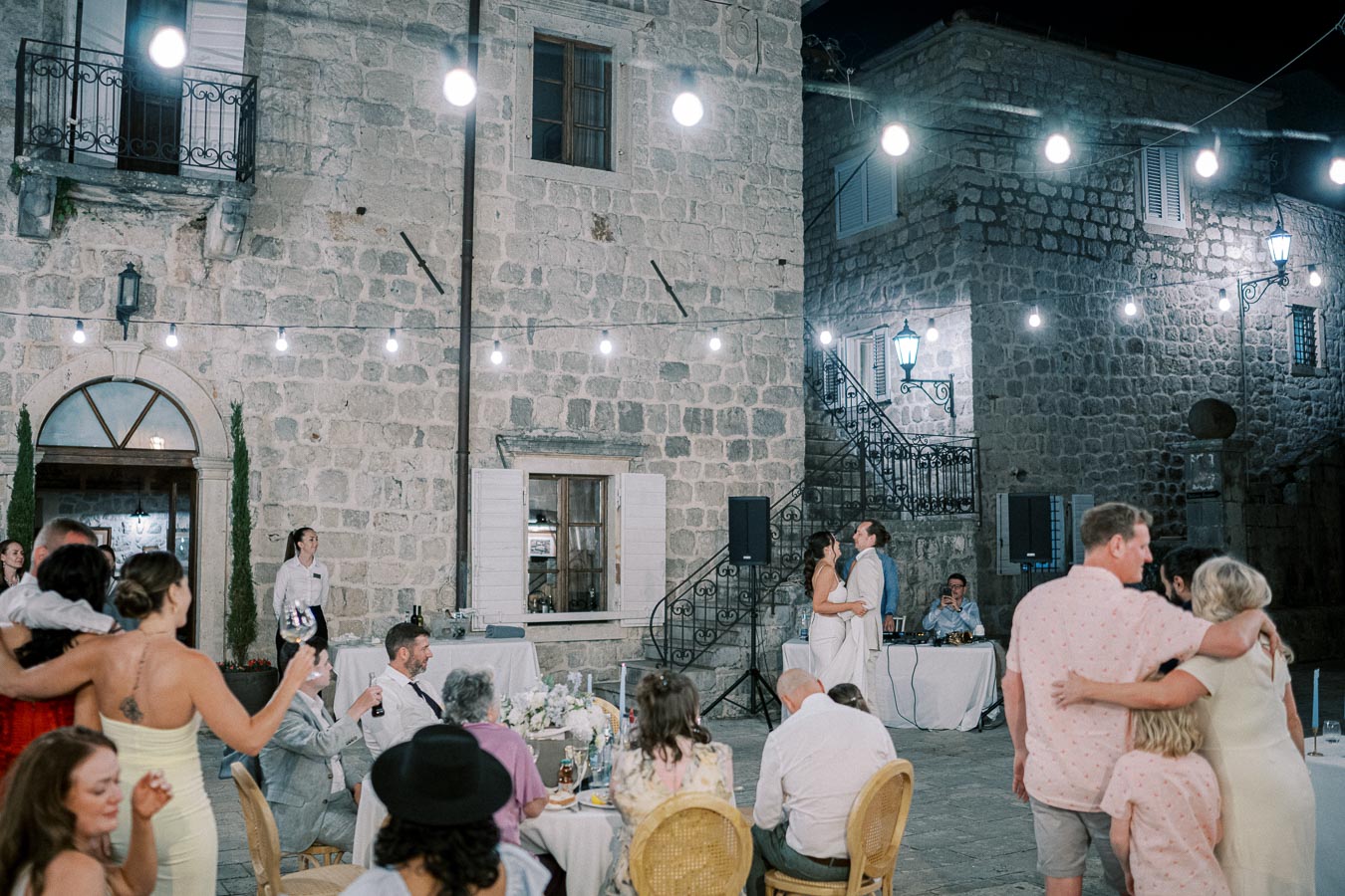 Outdoor evening wedding reception in a charming rustic courtyard, with a couple dancing under string lights surrounded by guests celebrating.