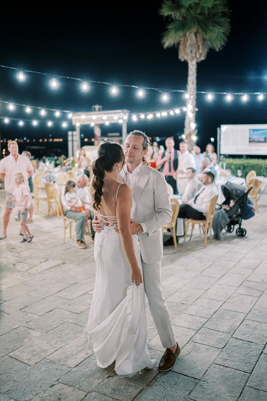 A couple shares a first dance under string lights at an outdoor wedding reception, surrounded by guests seated at tables, with a palm tree and night sky backdrop.