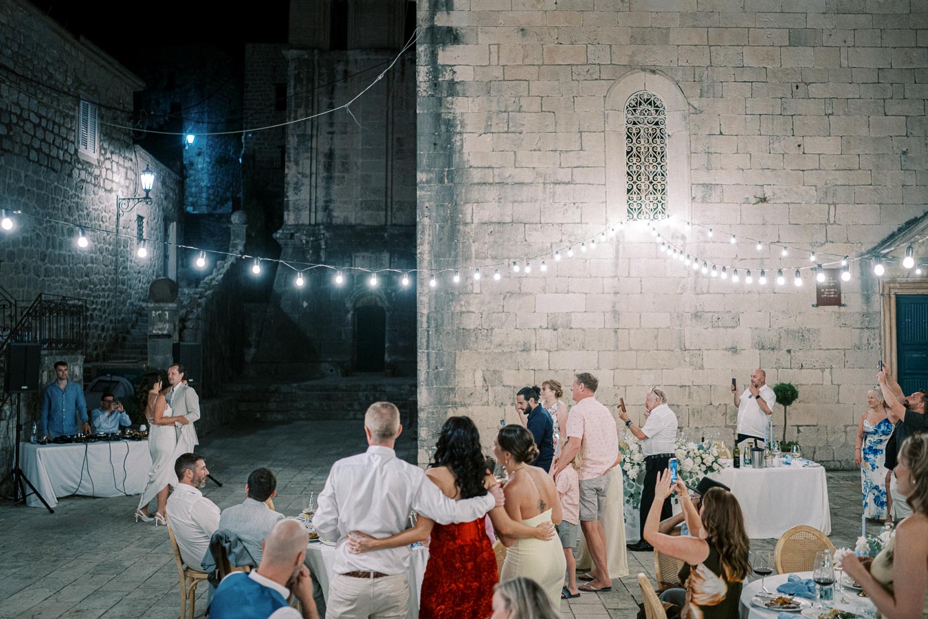 Outdoor wedding reception at night in a historic stone courtyard, featuring string lights, a DJ setup, and guests watching a couple's first dance.