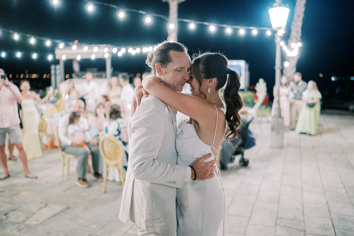 Romantic couple dancing closely under twinkling string lights at an outdoor evening event, surrounded by seated guests on a stone patio.