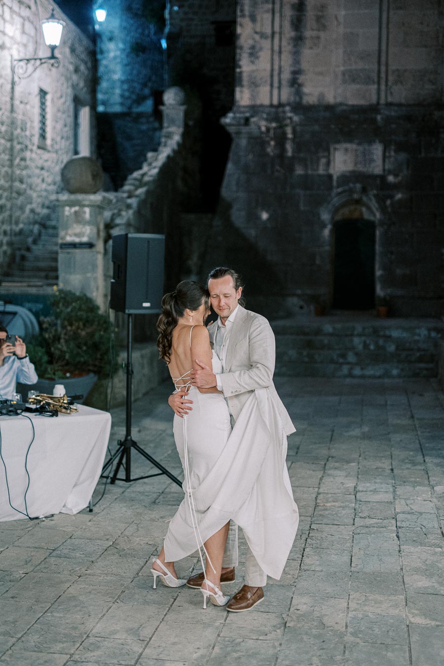 Couple dancing gracefully in an outdoor evening wedding setting, surrounded by historic stone architecture, capturing a romantic moment under soft ambient lighting.