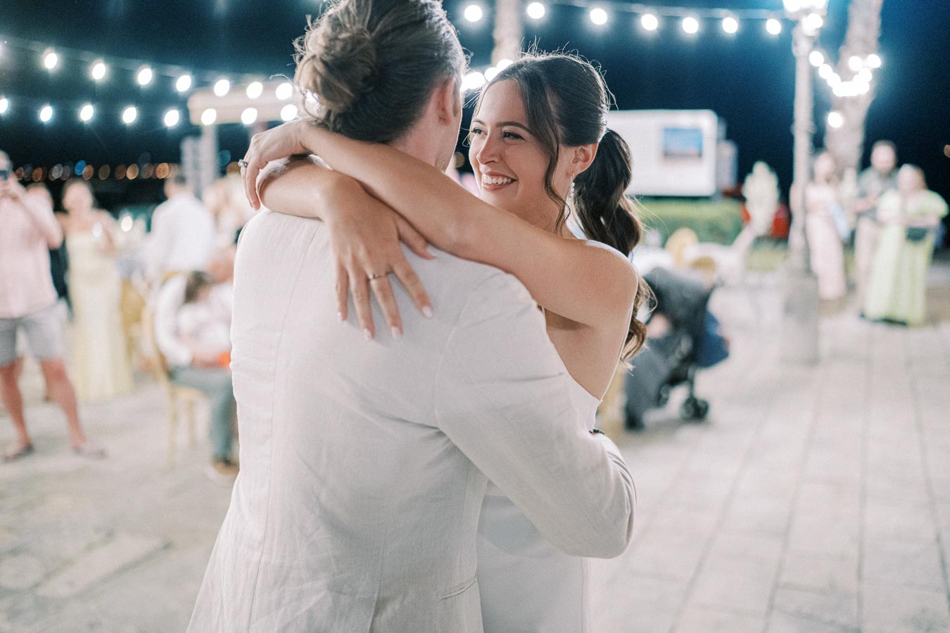 Happy couple sharing a romantic first dance at a beautifully lit outdoor wedding reception with guests in the background.