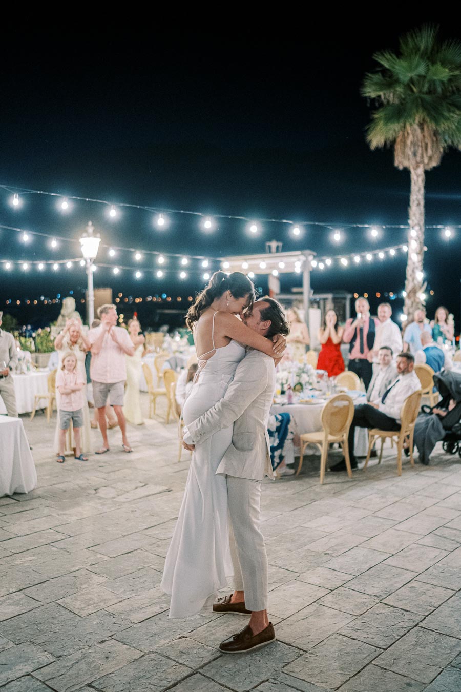 Romantic outdoor wedding dance under string lights, bride embraced by groom with guests celebrating in the background.