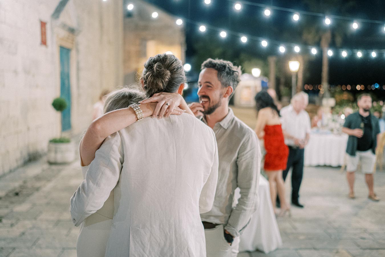 A joyful wedding reception scene with string lights overhead, showing a warmly embraced couple in white attire and a smiling man in a gray shirt. Guests mingle in the background under the soft evening glow, celebrating outdoors.