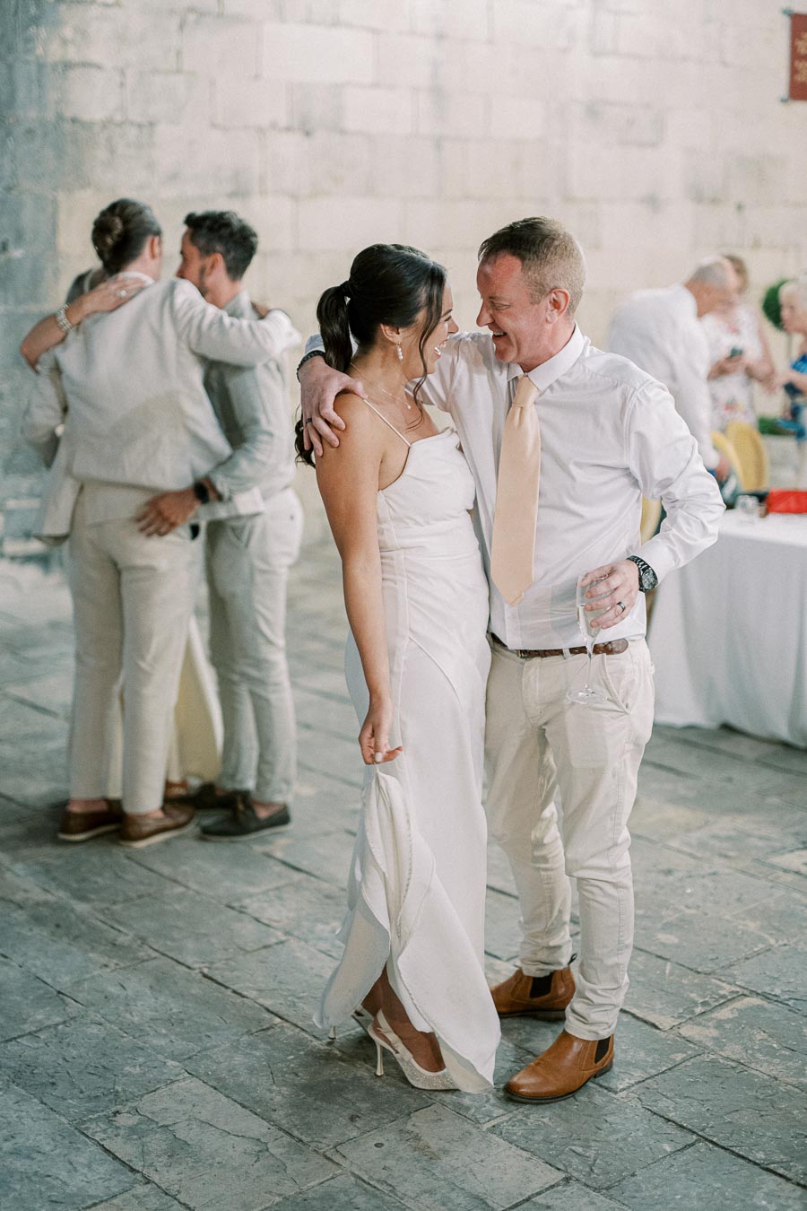 A couple dressed in elegant white attire shares a joyful moment at a wedding reception, with another couple embracing in the background.