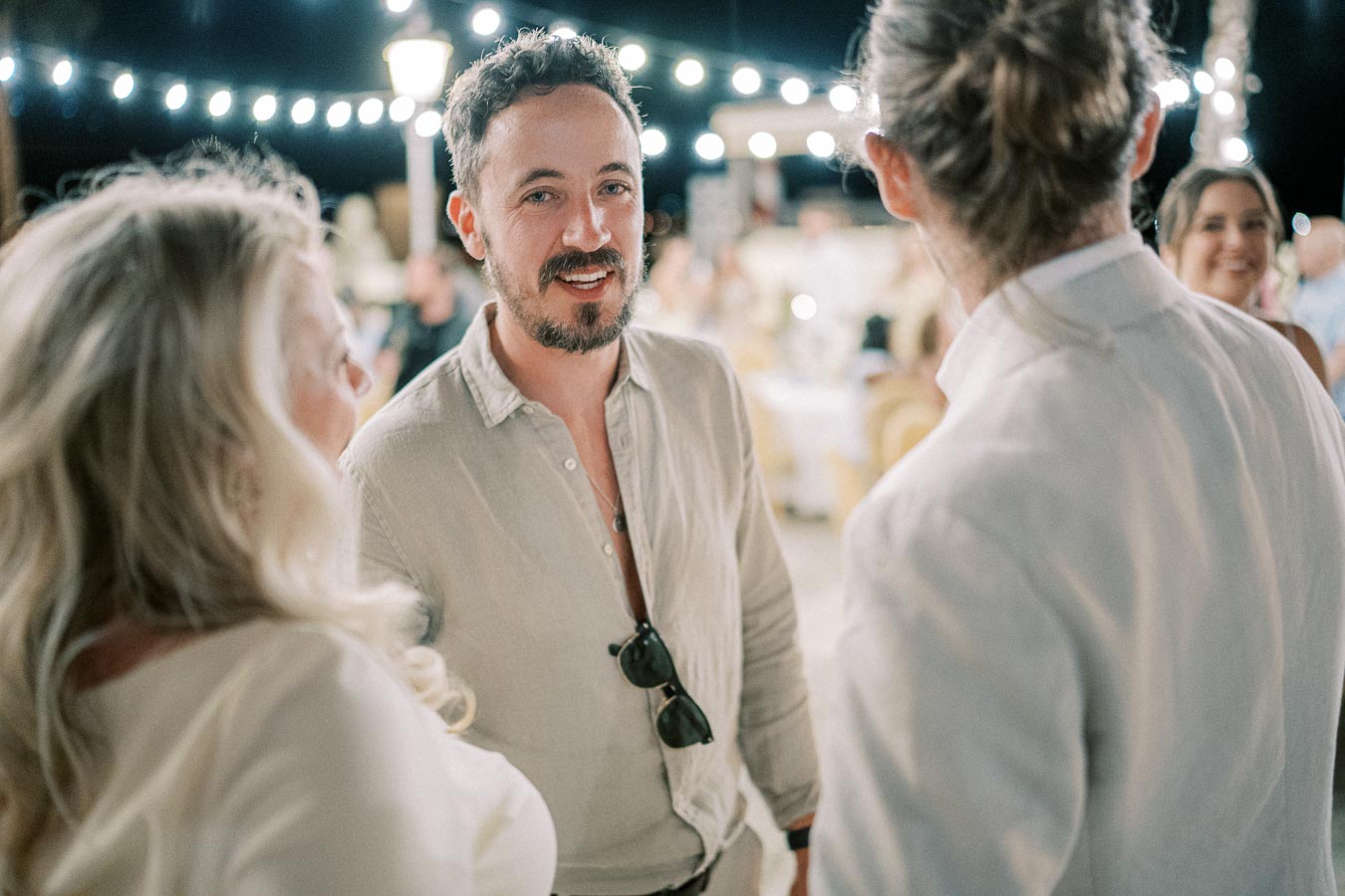 A lively outdoor evening gathering with people socializing, featuring a man in casual attire engaged in conversation, surrounded by twinkling string lights and a festive ambiance.