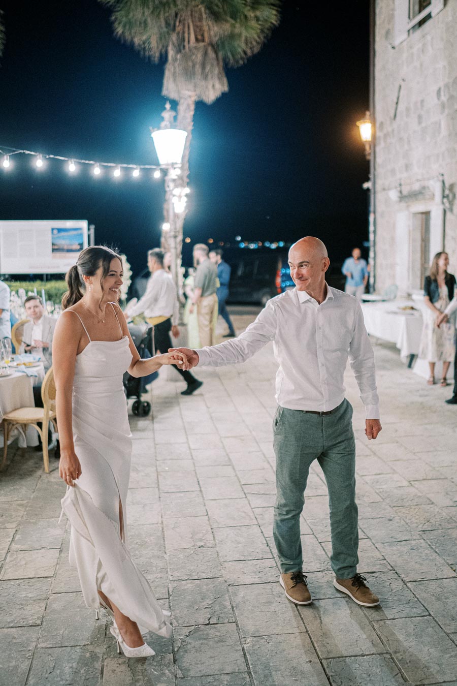 A joyful couple dancing at an outdoor evening event, surrounded by string lights and a scenic background, with people socializing nearby.