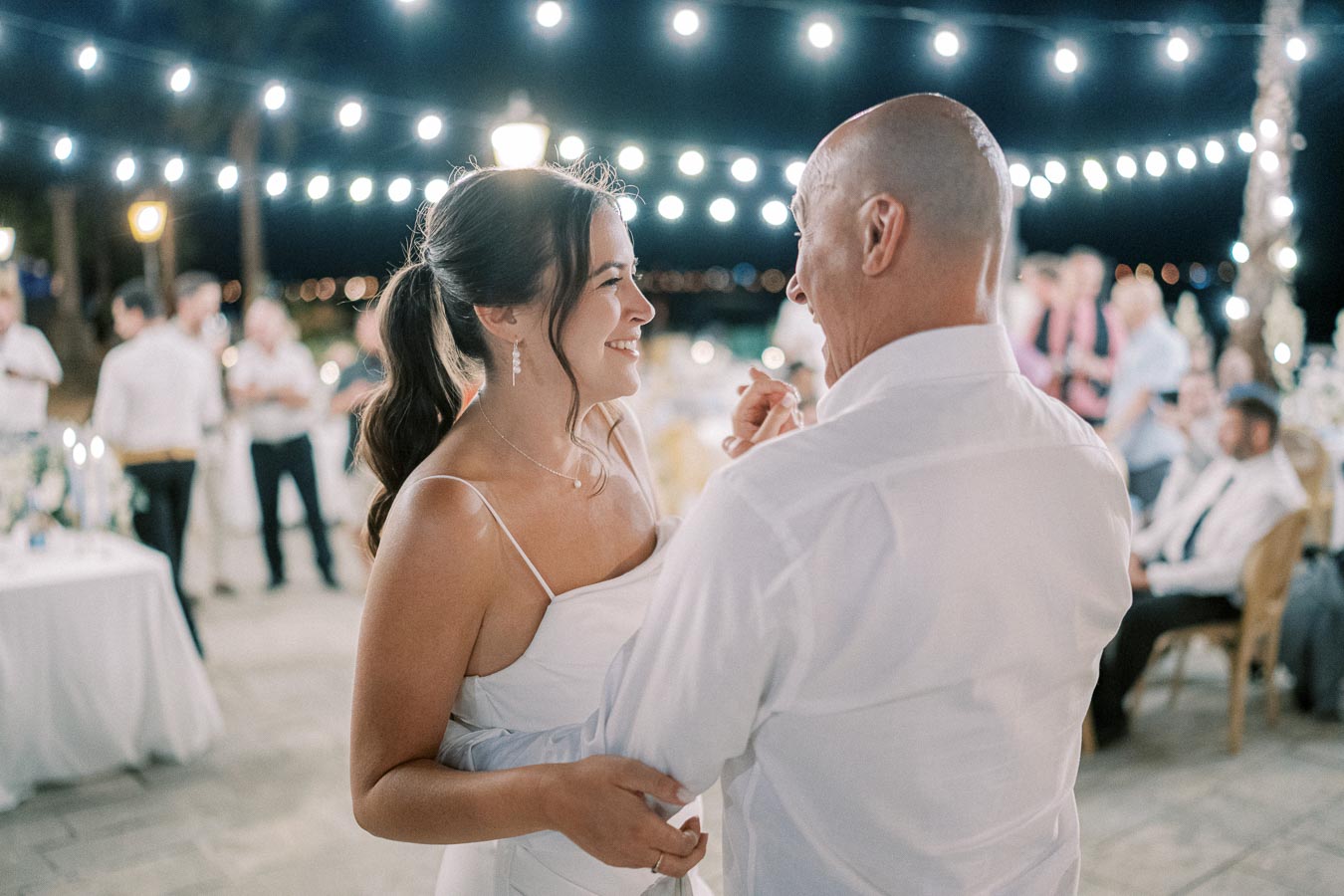 Father and daughter share a joyful dance at an outdoor evening wedding reception, illuminated by string lights.