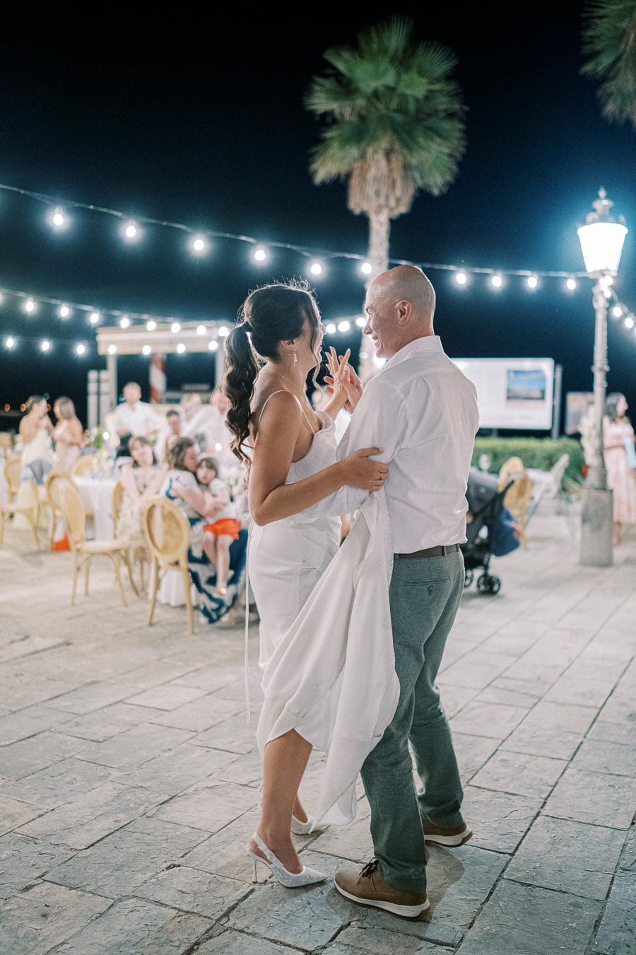 Couple dancing under string lights at an outdoor evening wedding reception, surrounded by guests seated at decorated tables.