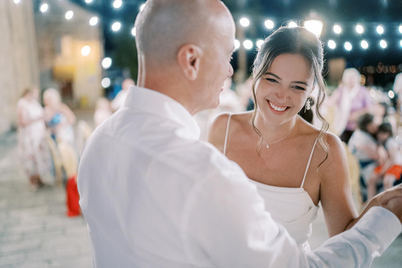 A bride joyfully dancing with a guest under twinkling string lights at an outdoor wedding reception, surrounded by guests in festive attire.