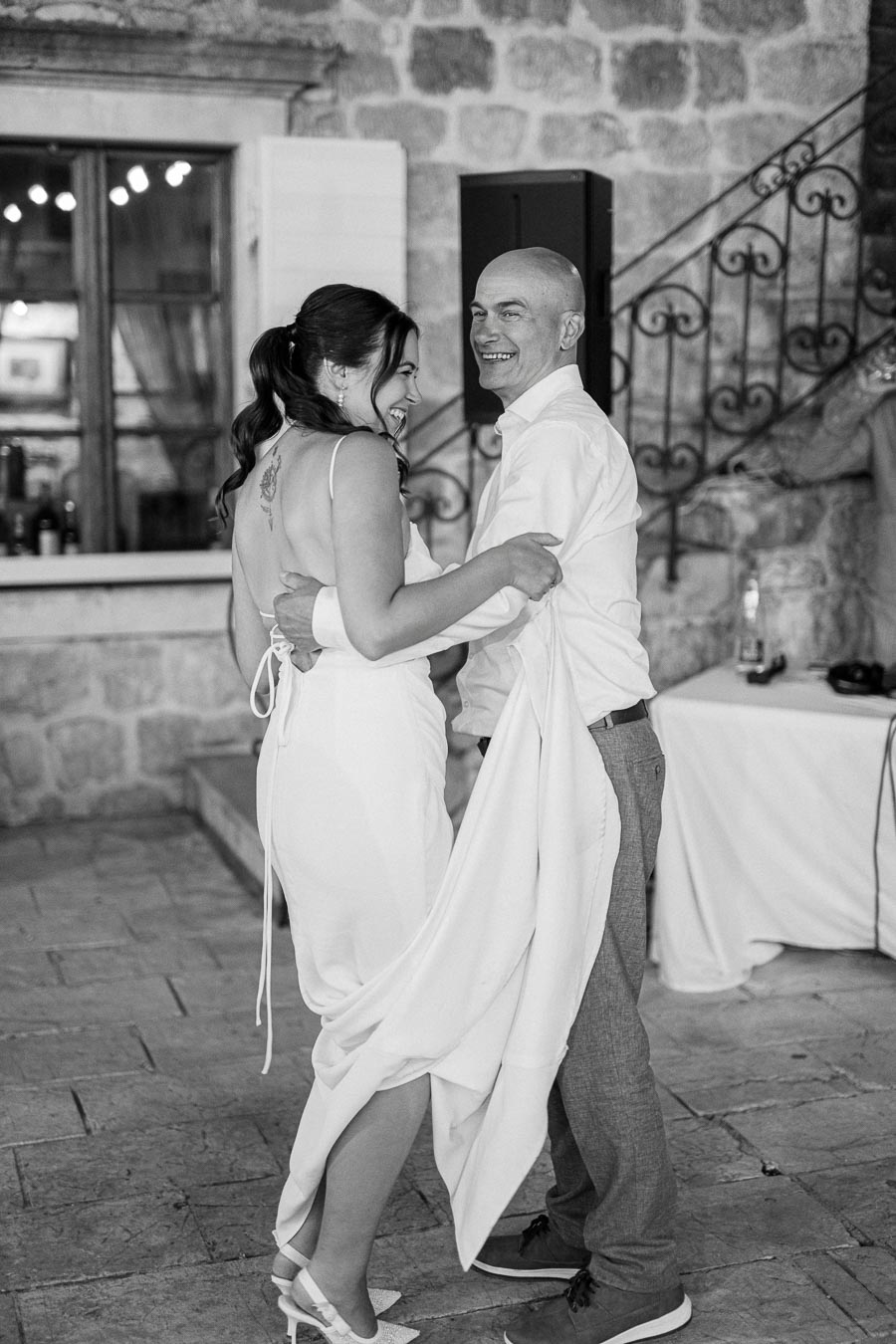 A couple joyfully dancing in a rustic setting, with stone walls in the background. The woman is wearing a white dress and heels, and the man in a light shirt and trousers. The mood captures a candid and happy moment.