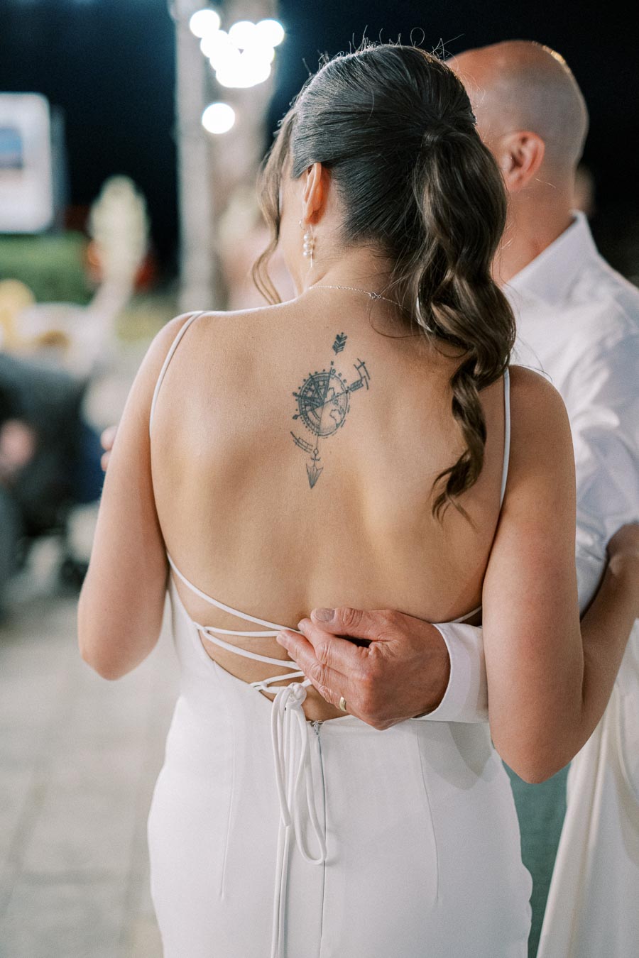 Bride wearing a backless white dress with a compass-themed tattoo dancing with a partner at a nighttime outdoor wedding reception.
