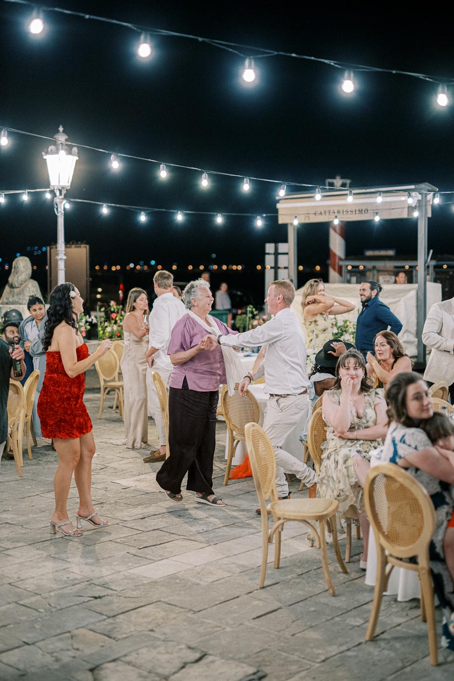 Outdoor evening event with people dancing and socializing under string lights. Guests in vibrant attire appear joyful, enjoying a festive atmosphere with tables and chairs set against a night backdrop.