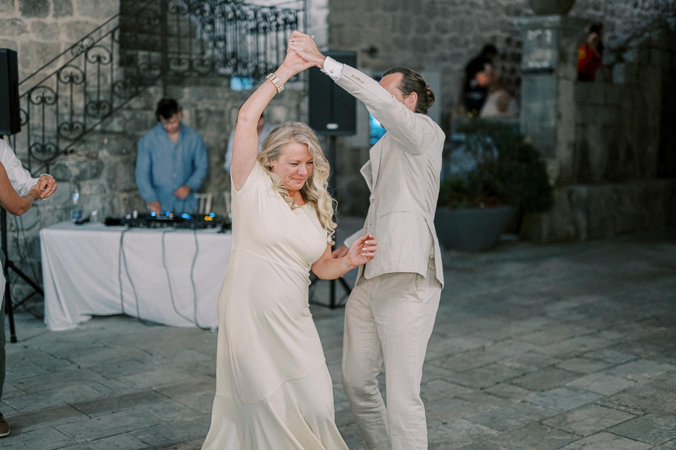 Couple joyfully dancing at a wedding reception with a DJ in the background, set in a rustic stone venue.