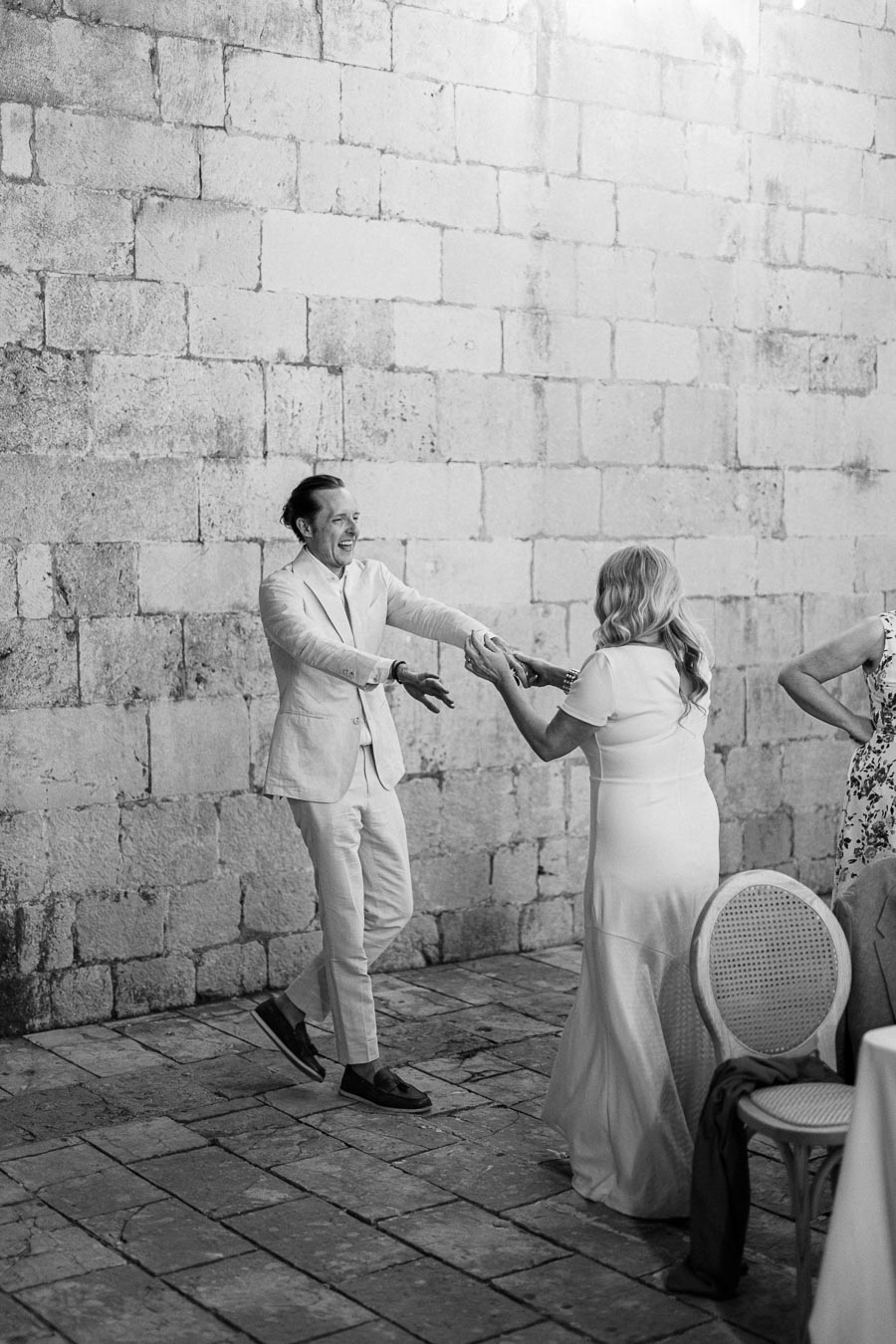 A black and white photo of a couple joyfully dancing on a cobblestone floor next to a rustic stone wall, capturing a lively celebration atmosphere.