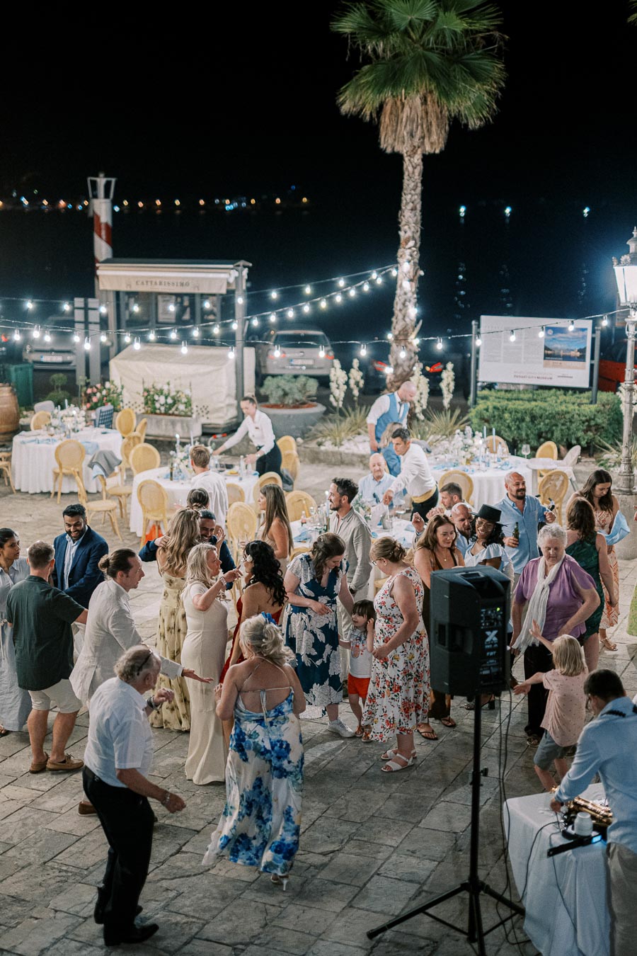 Outdoor evening wedding reception with guests dancing under string lights by the sea, surrounded by palm trees and elegantly decorated tables.