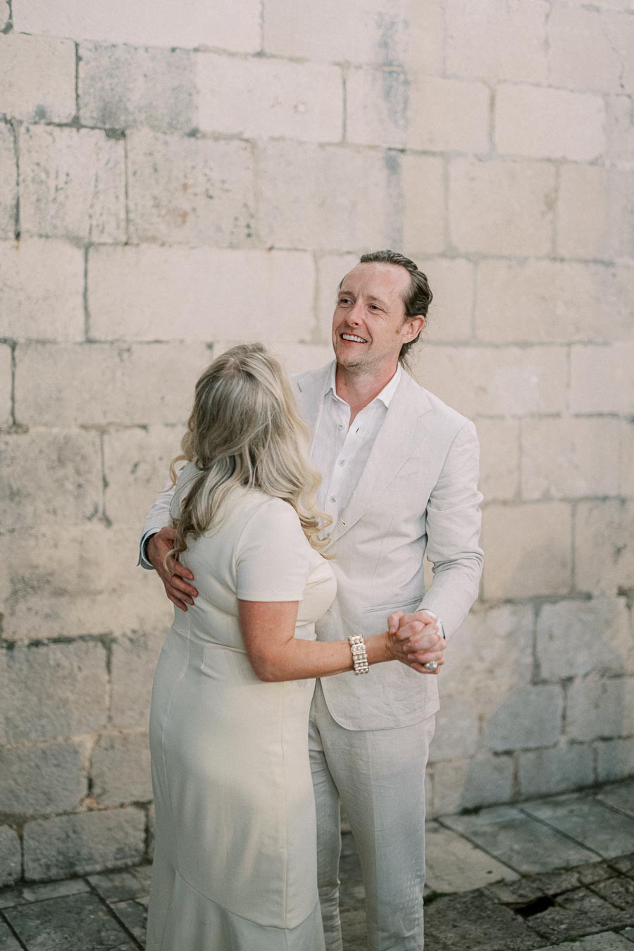 A couple in elegant attire dancing happily in front of a rustic stone wall. The man wears a light suit, while the woman is in a cream dress, creating a romantic and timeless scene.