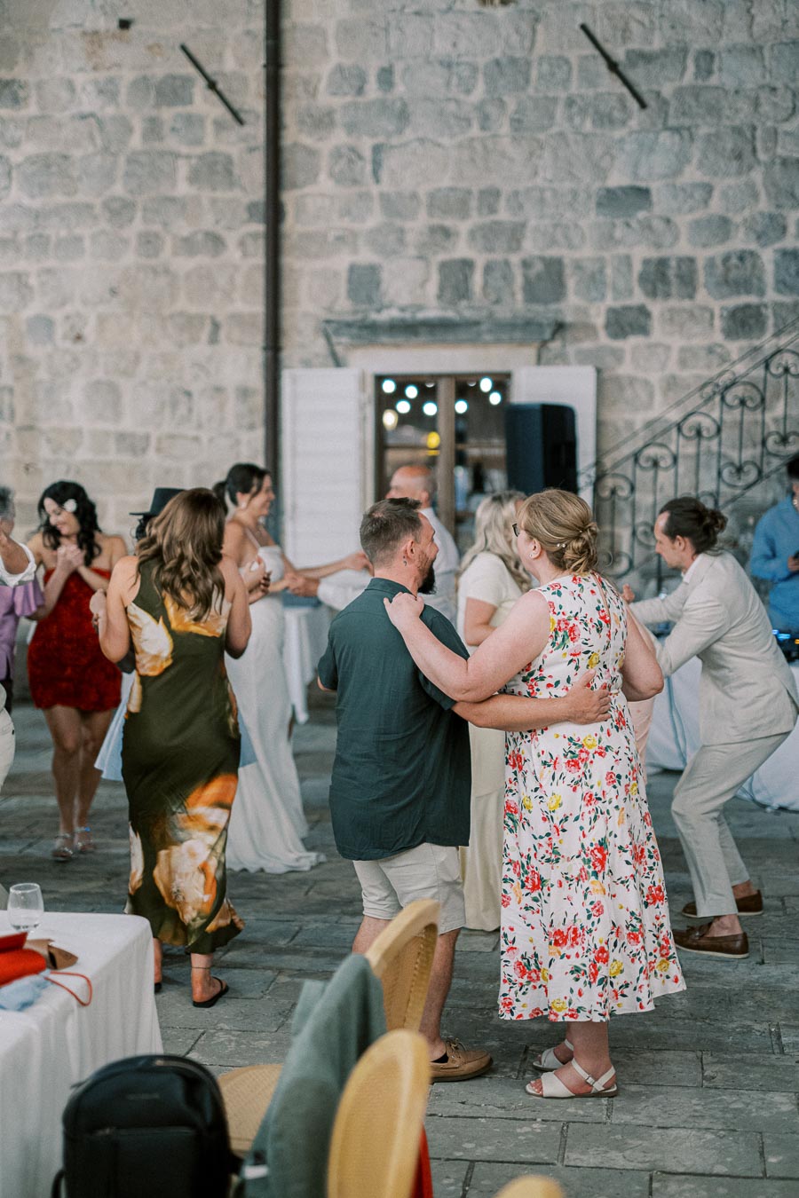A group of people dancing at an outdoor celebration against a stone wall backdrop, featuring a variety of colorful outfits and a lively atmosphere.