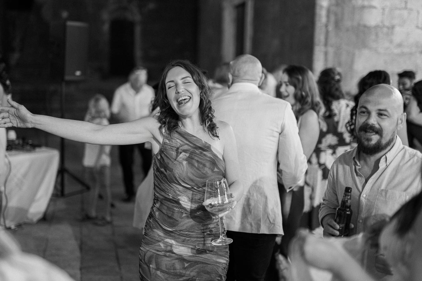 Black and white photo of a joyful woman singing at a party, holding a wine glass, with guests mingling in the background.