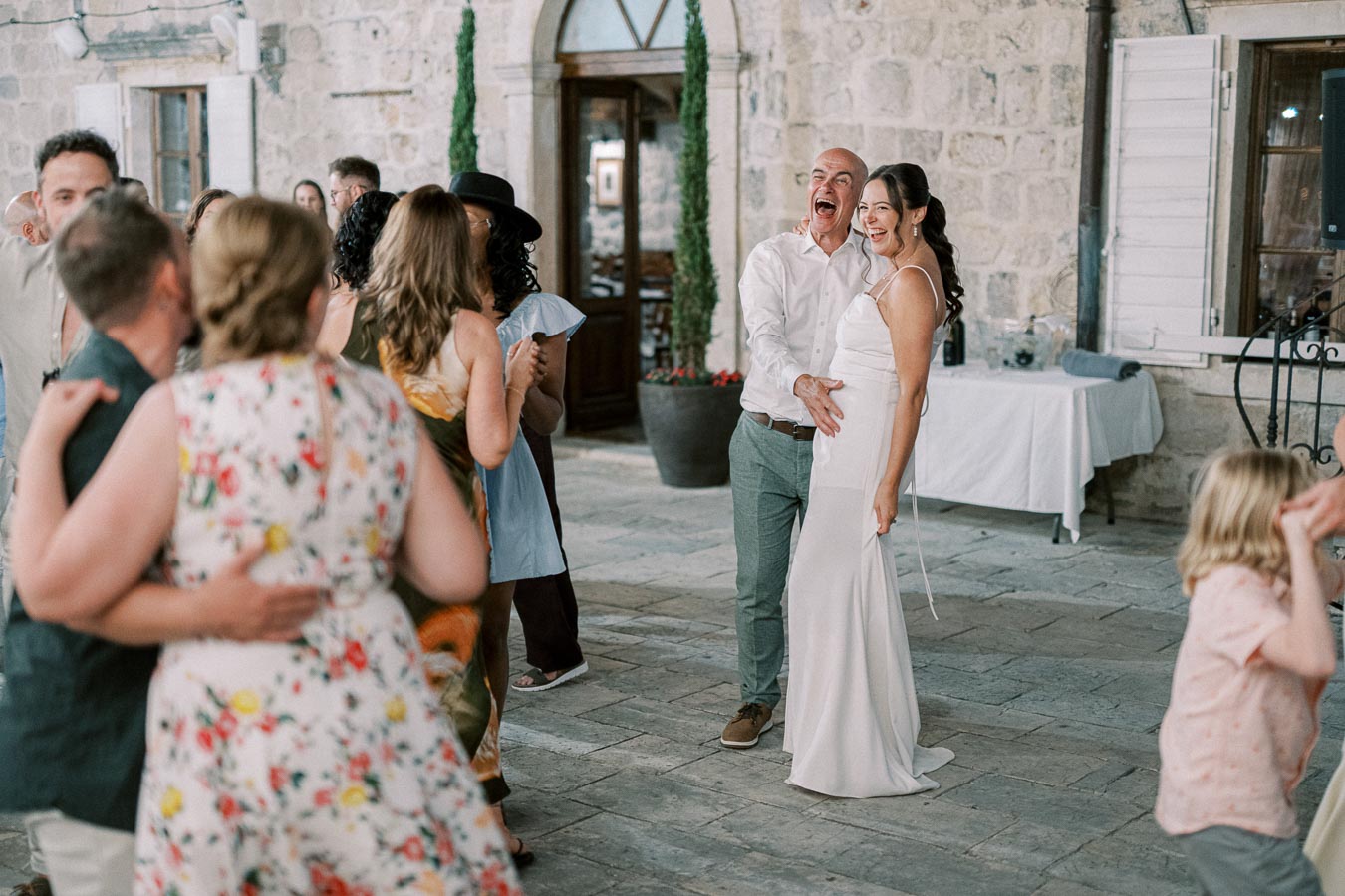 A joyful wedding reception with a bride in a white gown and a groom celebrating on a stone patio surrounded by guests dancing in colorful attire.