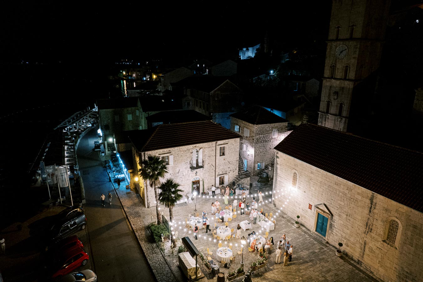 Aerial view of an elegant outdoor evening wedding reception in a historic stone courtyard, featuring round tables adorned with white linens and vibrant decorations, surrounded by string lights, with guests gathered around, creating a warm, festive atmosphere.