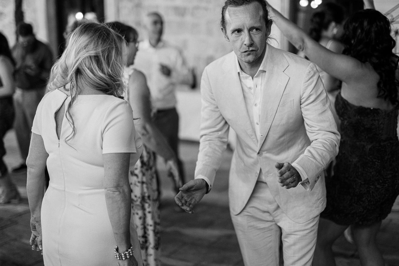 Black and white photo of a man in a light-colored suit and a woman in a dress dancing at a wedding reception, with other guests socializing in the background.