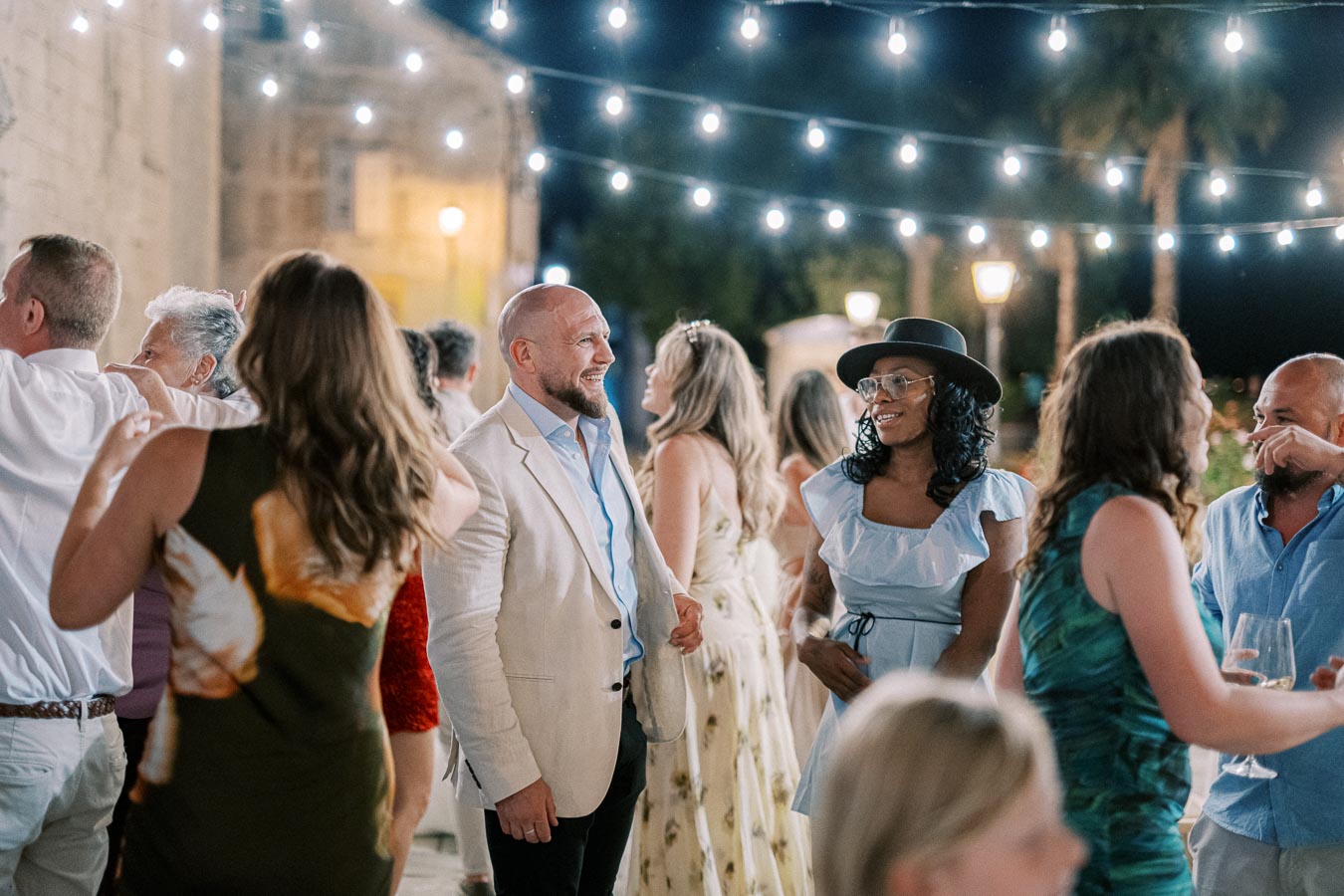 Group of people enjoying an evening outdoor party with string lights, socializing and smiling in elegant attire.