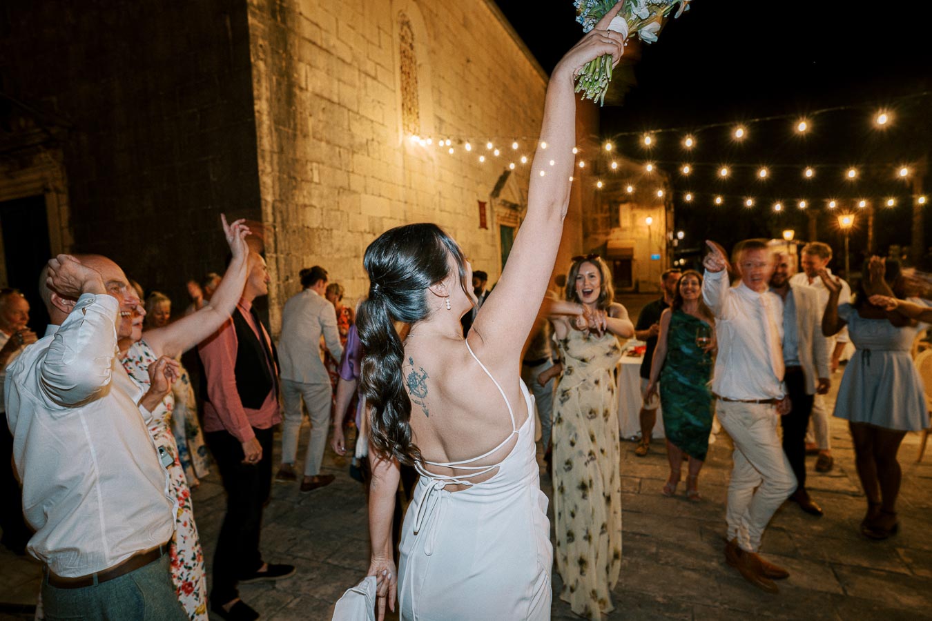 Bride tossing bouquet at night outdoor wedding celebration, guests cheering under string lights.