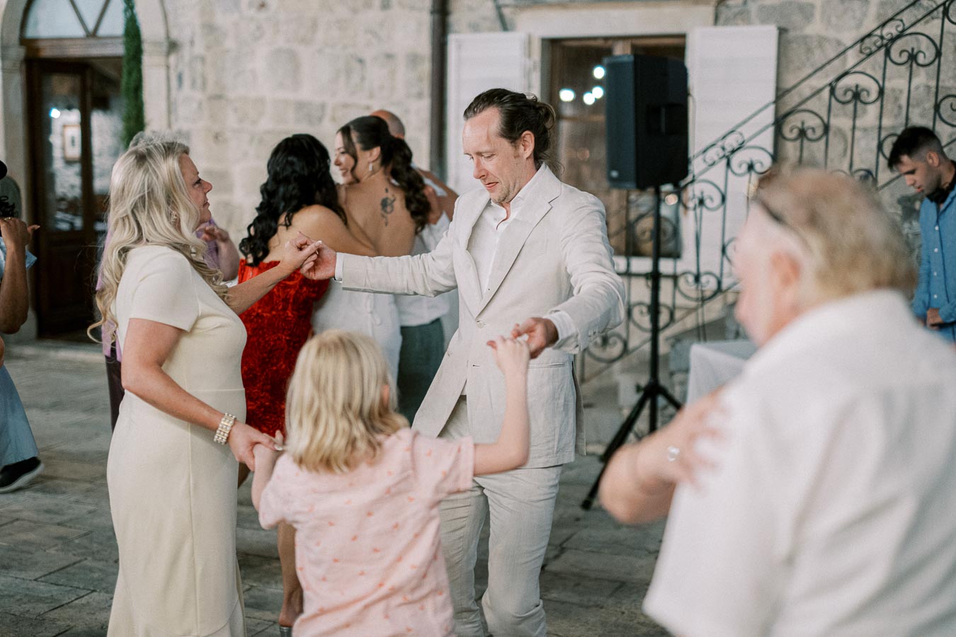 A joyful group of people dancing together at an outdoor event, with a man in a white suit holding hands with a woman in a white dress and a child, surrounded by others in a lively celebration atmosphere.