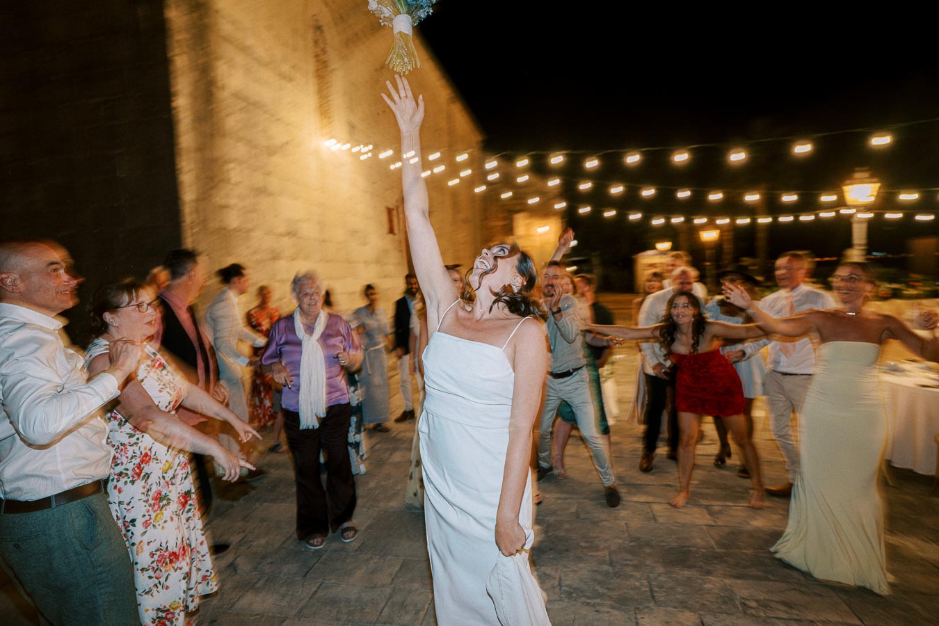 Wedding reception bouquet toss with bride in white dress, reaching overhead surrounded by smiling guests under string lights at night.