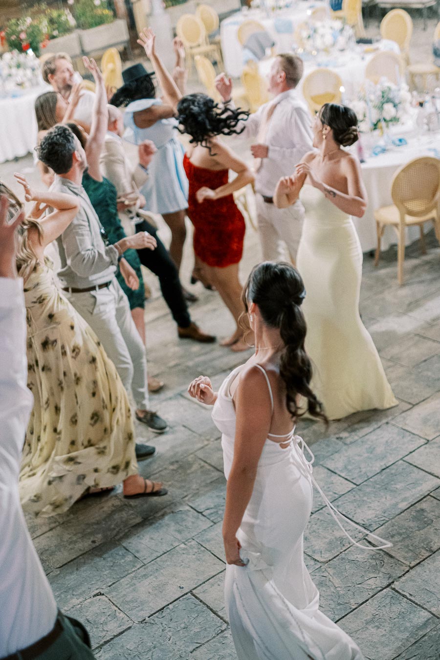A group of people dancing energetically at an indoor wedding reception, with elegantly set tables and floral decorations in the background.