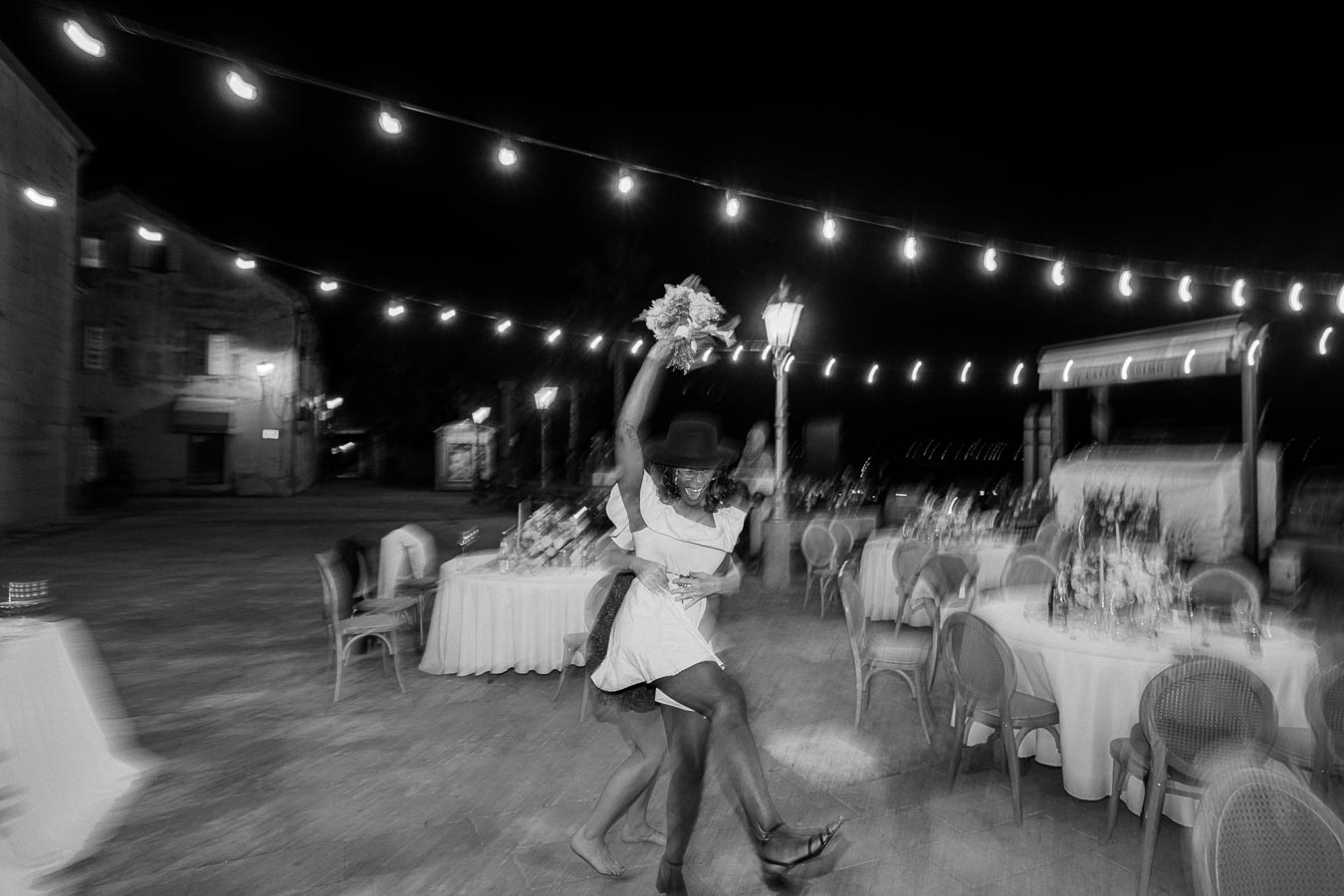 A joyous couple dancing at an outdoor evening wedding reception, illuminated by string lights and surrounded by elegantly set tables.