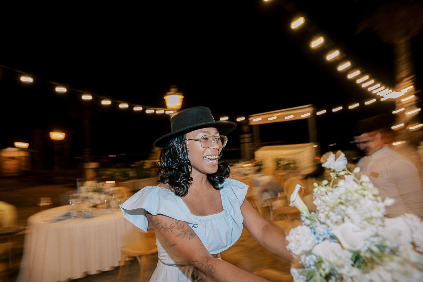 A woman wearing a black hat and glasses, smiling while holding a bouquet of flowers at an outdoor evening event with string lights in the background.