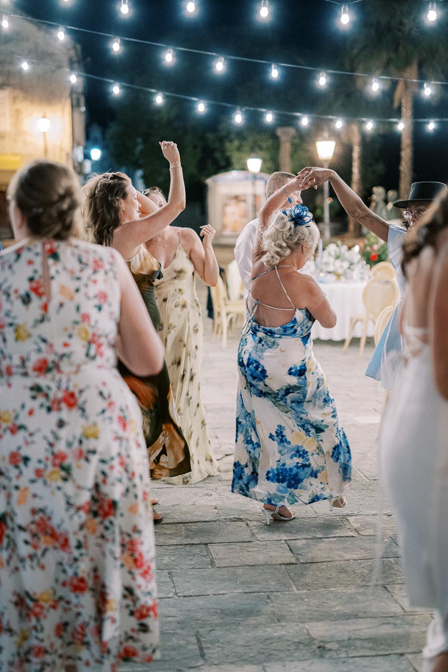 People dancing joyfully under string lights at an outdoor evening event, wearing floral dresses and celebrating in a festive atmosphere.