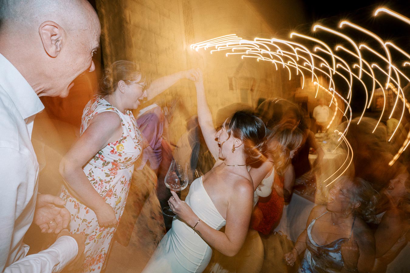 A group of people dancing energetically at an evening event, with decorative lights creating a dynamic swirl effect.